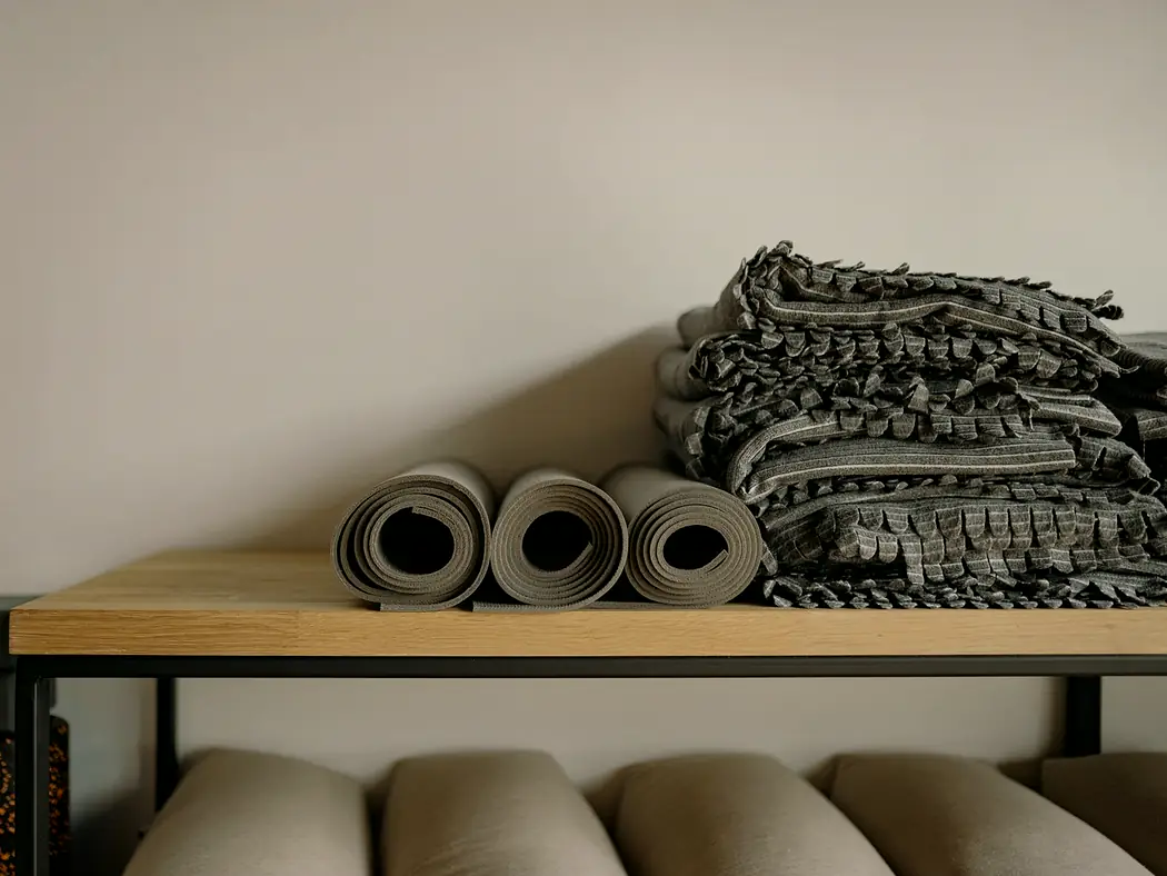 Rolled yoga mats, folded blankets and bolsters stacked on a wooden shelf.